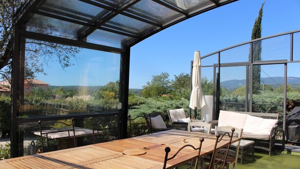 A covered patio with a wooden table and chairs, a white sofa, and a view of the surrounding landscape. - House Les Hortensias (MONTAUROUX)
