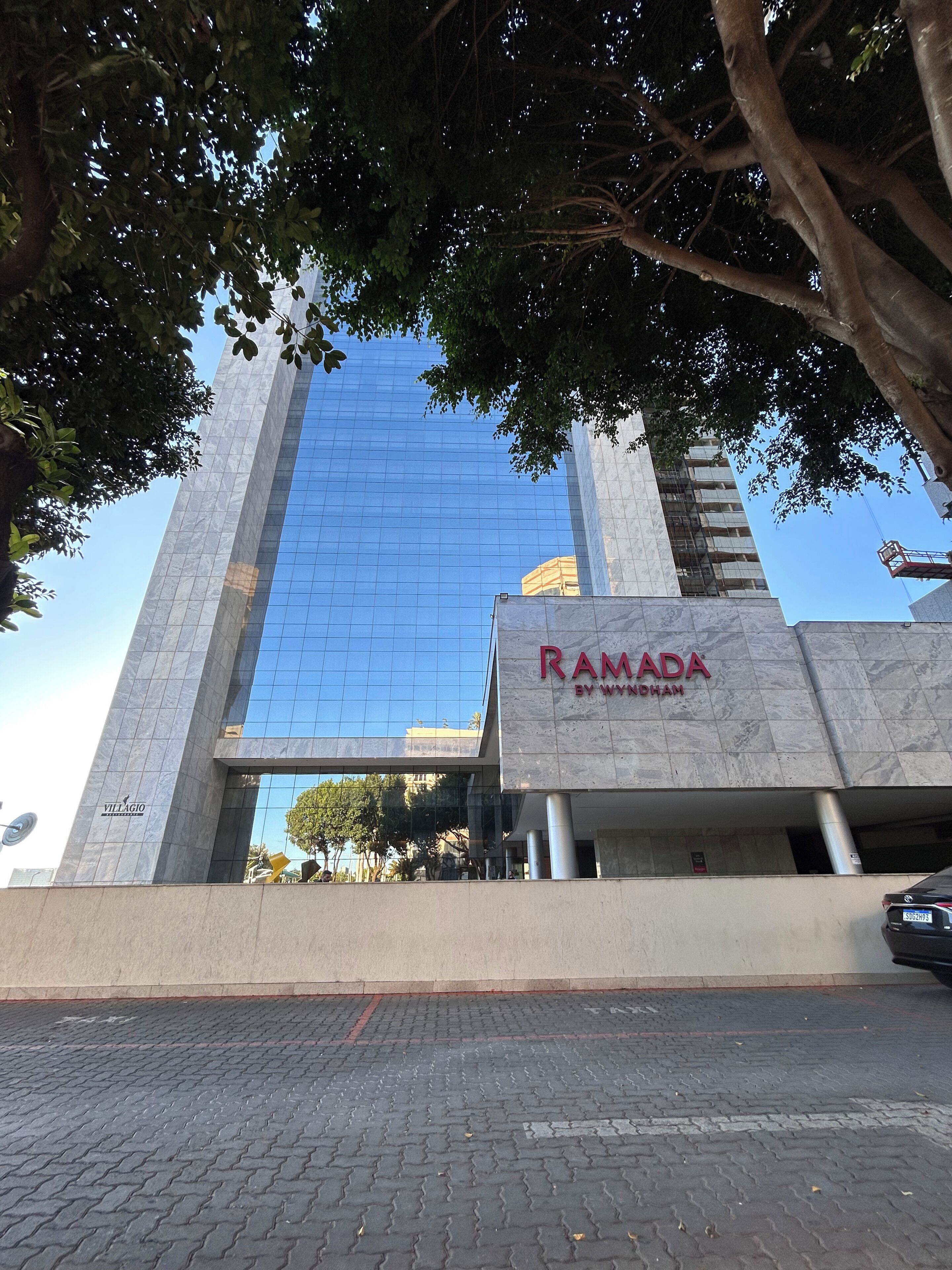Ein modernes Gebäude mit Glasfassade, einem gut sichtbaren Schild für 'RAMADA BY WYNDHAM' und einem gepflasterten Eingangsbereich.