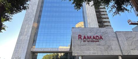 Ein modernes Gebäude mit Glasfassade, einem gut sichtbaren Schild für 'RAMADA BY WYNDHAM' und einem gepflasterten Eingangsbereich.
