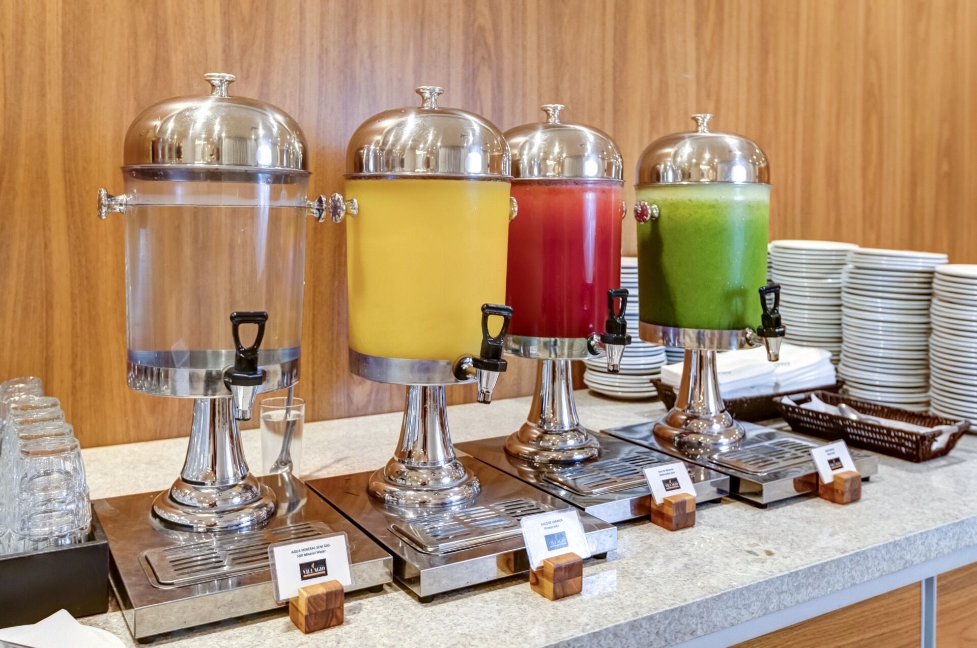 A beverage station with four dispensers, each with a different colored drink, and a stack of plates in the background.