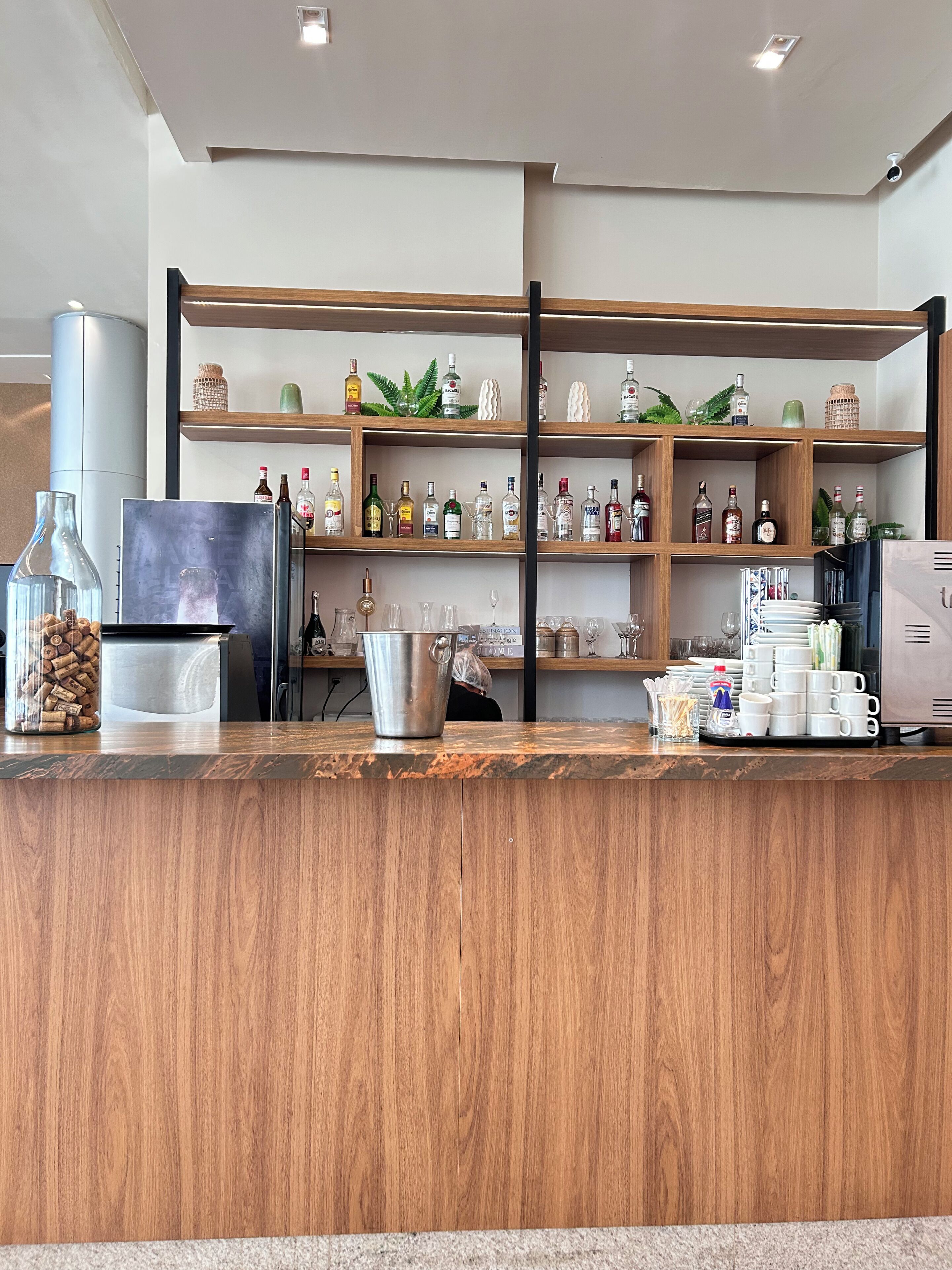 Una zona de bar con estantes de madera que exhiben botellas y vasos, una jarra de metal y una máquina de café.