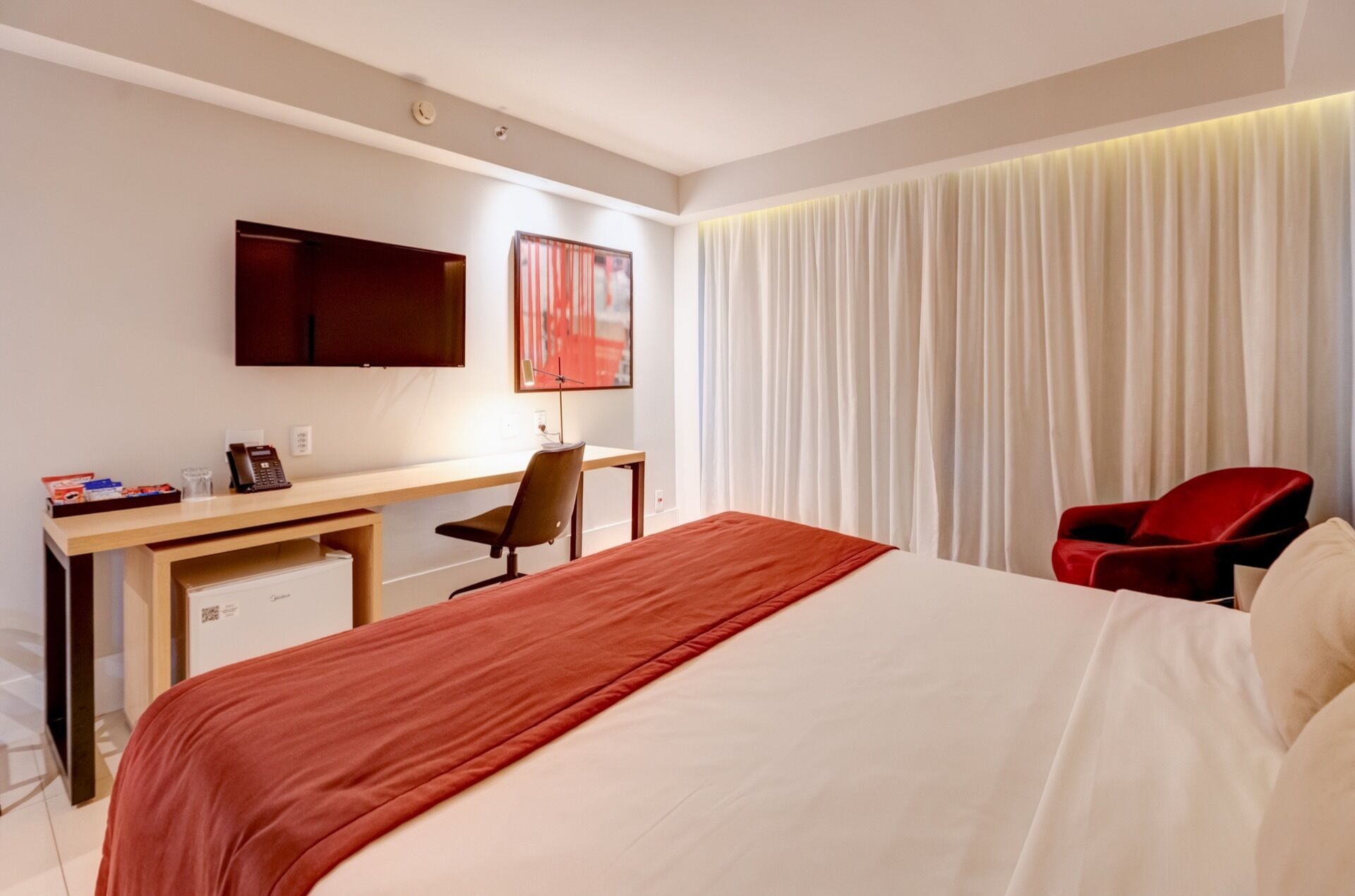A hotel room with a bed, a desk, a chair, a TV, and a window with curtains.