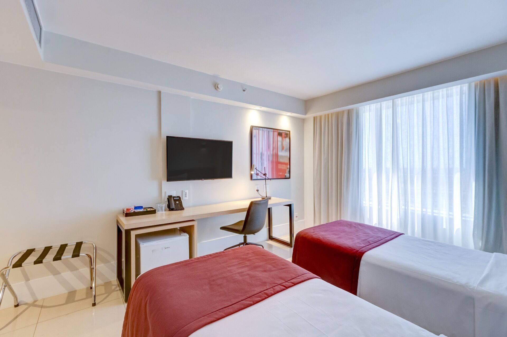 Une chambre d’hôtel comprenant un lit, un bureau avec une chaise, une télévision et une fenêtre avec des rideaux.