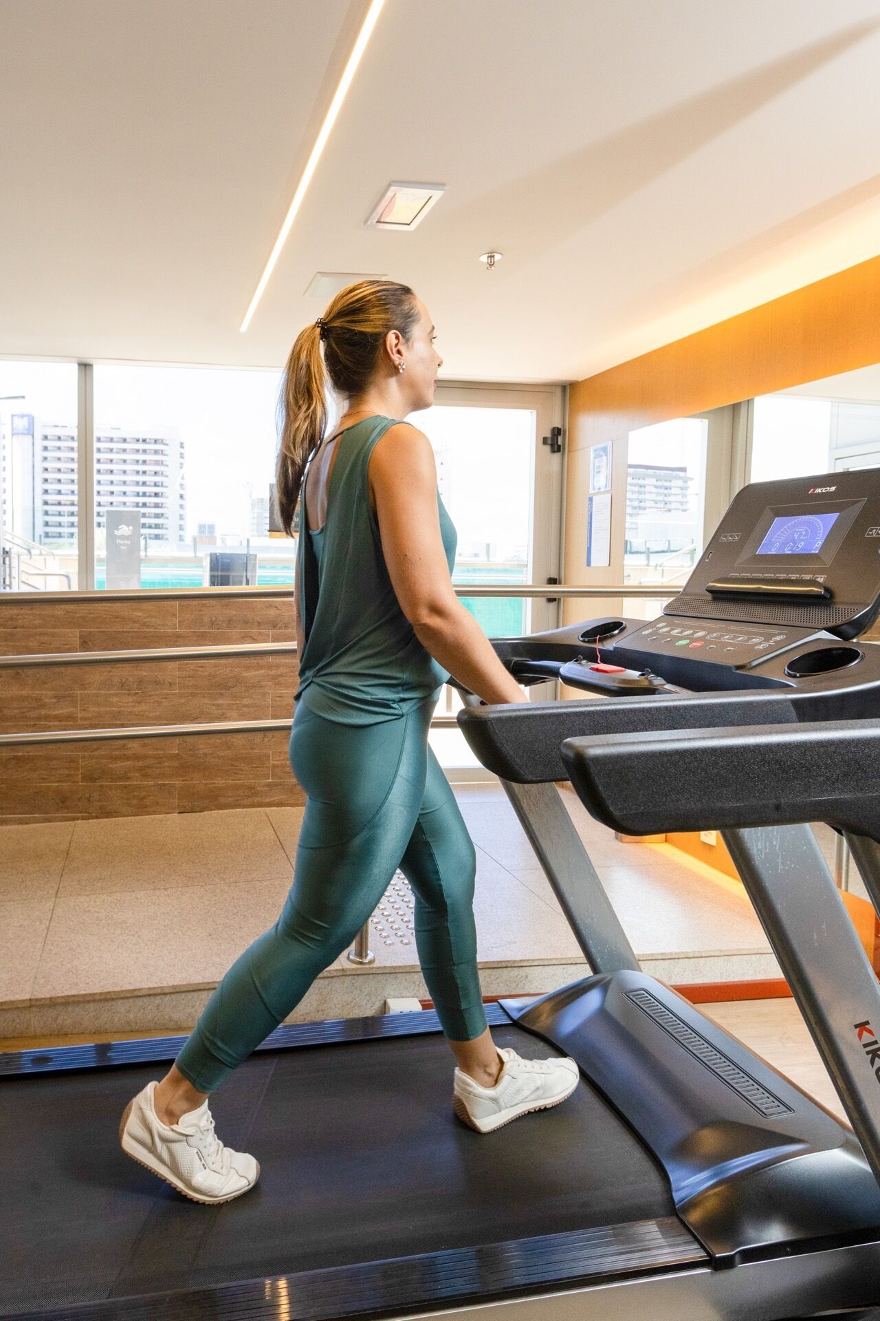 A woman walking on a treadmill in a gym with a city view.