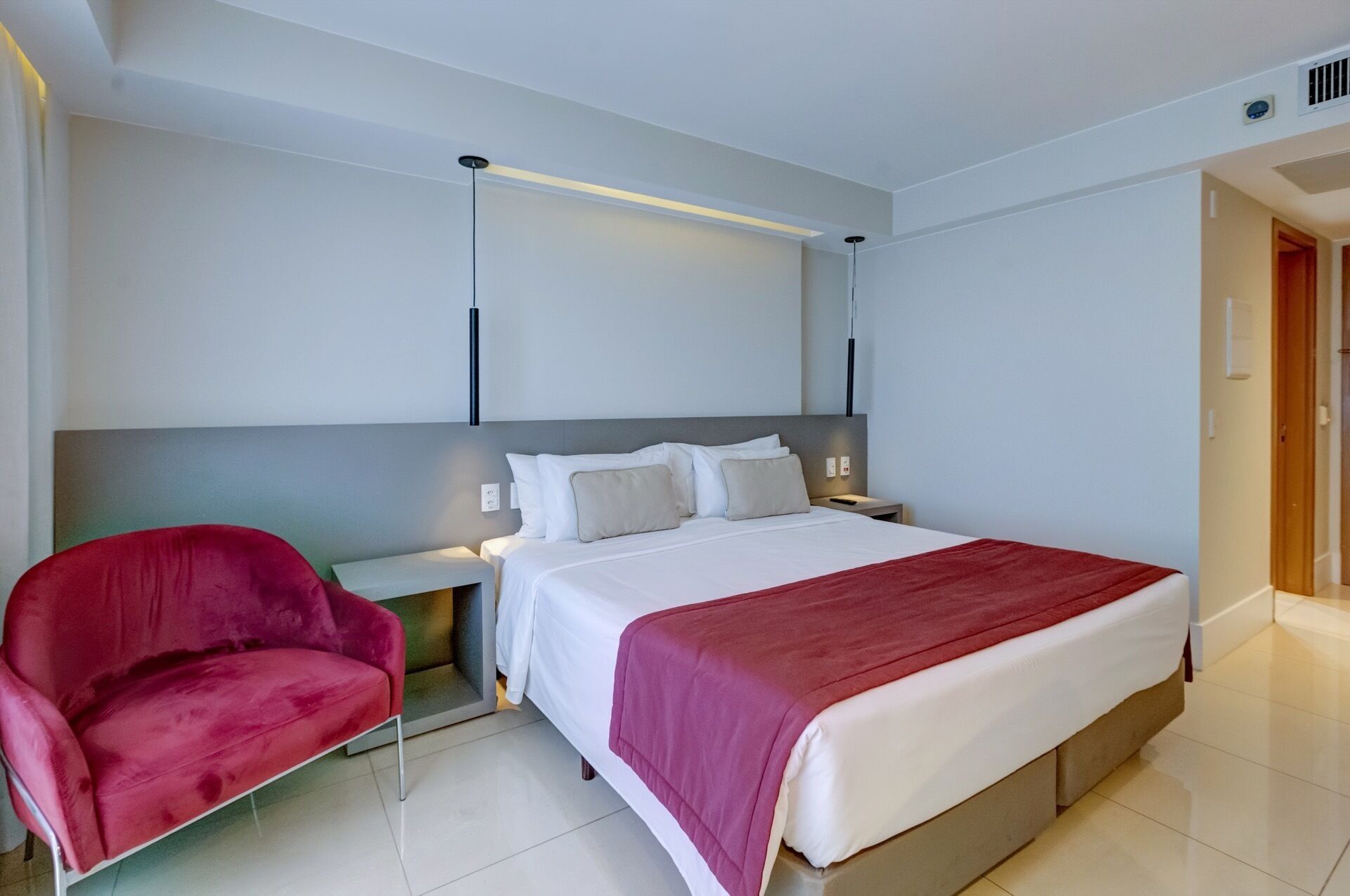 Une chambre d’hôtel avec un lit, une chaise rouge, une table de chevet et un luminaire fixé au mur.