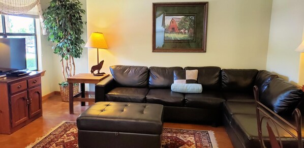 A living room with a leather sofa, ottoman, wooden side table with a lamp, a framed picture on the wall, and a television on a wooden cabinet.