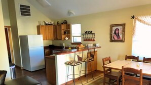 Cocina compacta con armarios de madera, un refrigerador y una barra con taburetes. Hay una mesa de comedor con sillas y un tapete estampado.