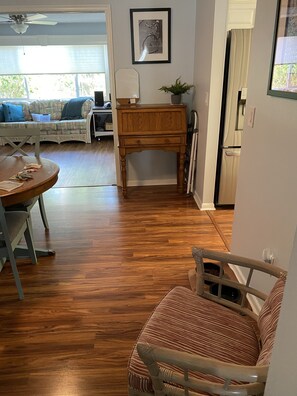 Een woonkamer met houten vloer, een eettafel met stoelen, een bank, een houten salontafel, een lijst met een foto aan de muur, en een deur die leidt naar een andere kamer.