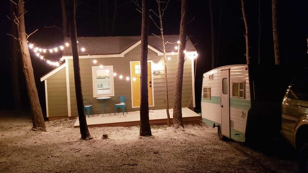 Eine kleine Holzhütte mit Veranda, Tisch und Stühlen, sowie ein Wohnwagen in der Nähe.