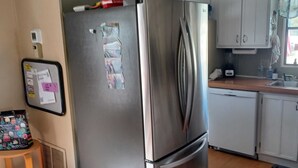 Una cocina con refrigerador de acero inoxidable, armarios blancos, un lavamanos pequeño y una ventana con cortina.