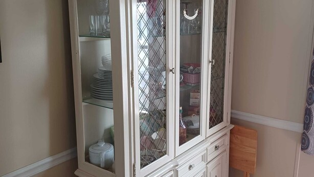 Un aparador blanco y ornamentado con varios cajones y puertas de cristal, que contiene distintos utensilios de cocina.