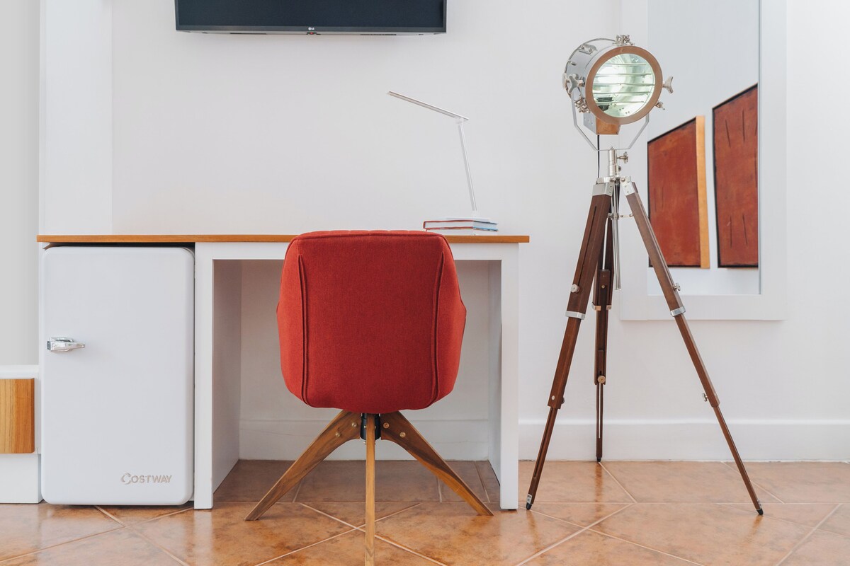 A room with a white desk, a red chair, a tripod with a camera, and a television on the wall.
