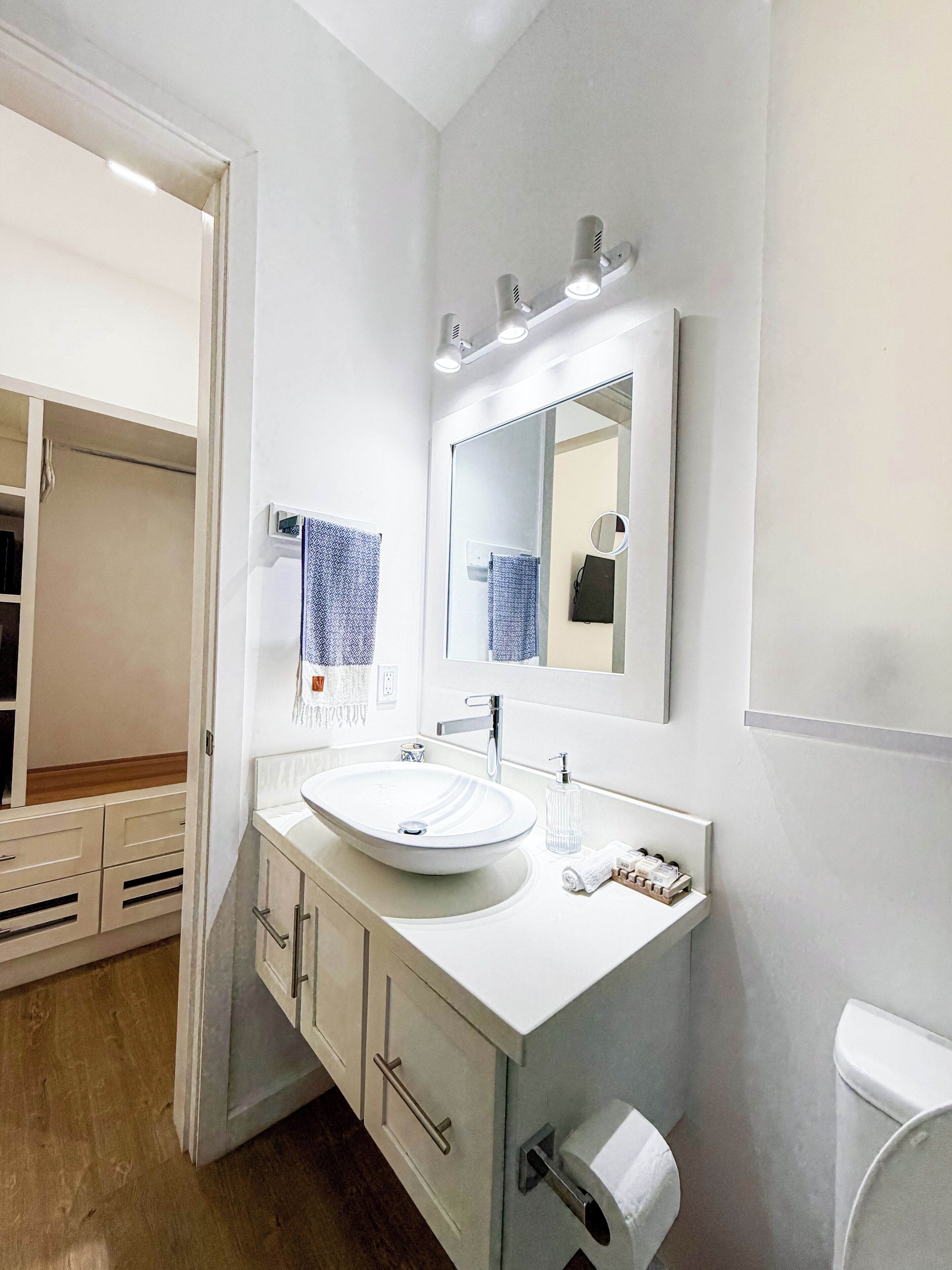 A modern bathroom with a white sink, vanity, and mirror. There is a toilet to the right and a wooden floor.