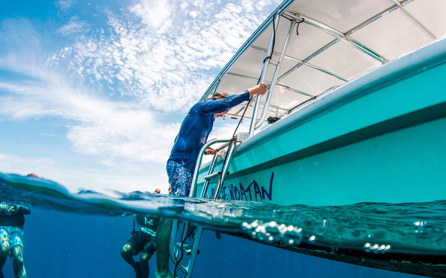 A diver ascending a ladder on a boat with the name "E. ROATAN" visible on its side.