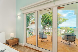 A room with a sliding glass door leading to an outdoor patio with wicker chairs and a view of the ocean. - Blue Roatan Resort (Roatan)
