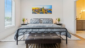 Ett sovrum med en säng, en bänk, en målning som föreställer en stadsbild och ett badrum som syns genom en öppen dörr.