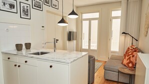 A modern kitchen with a marble countertop, a sink, and a built-in oven. There is a grey sofa with a wooden side table and a lamp.