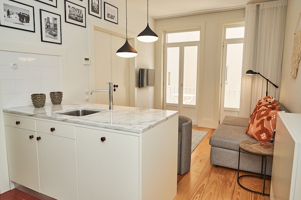 A modern kitchen with a marble countertop, a sink, and a built-in oven. There is a grey sofa with a wooden side table and a lamp.