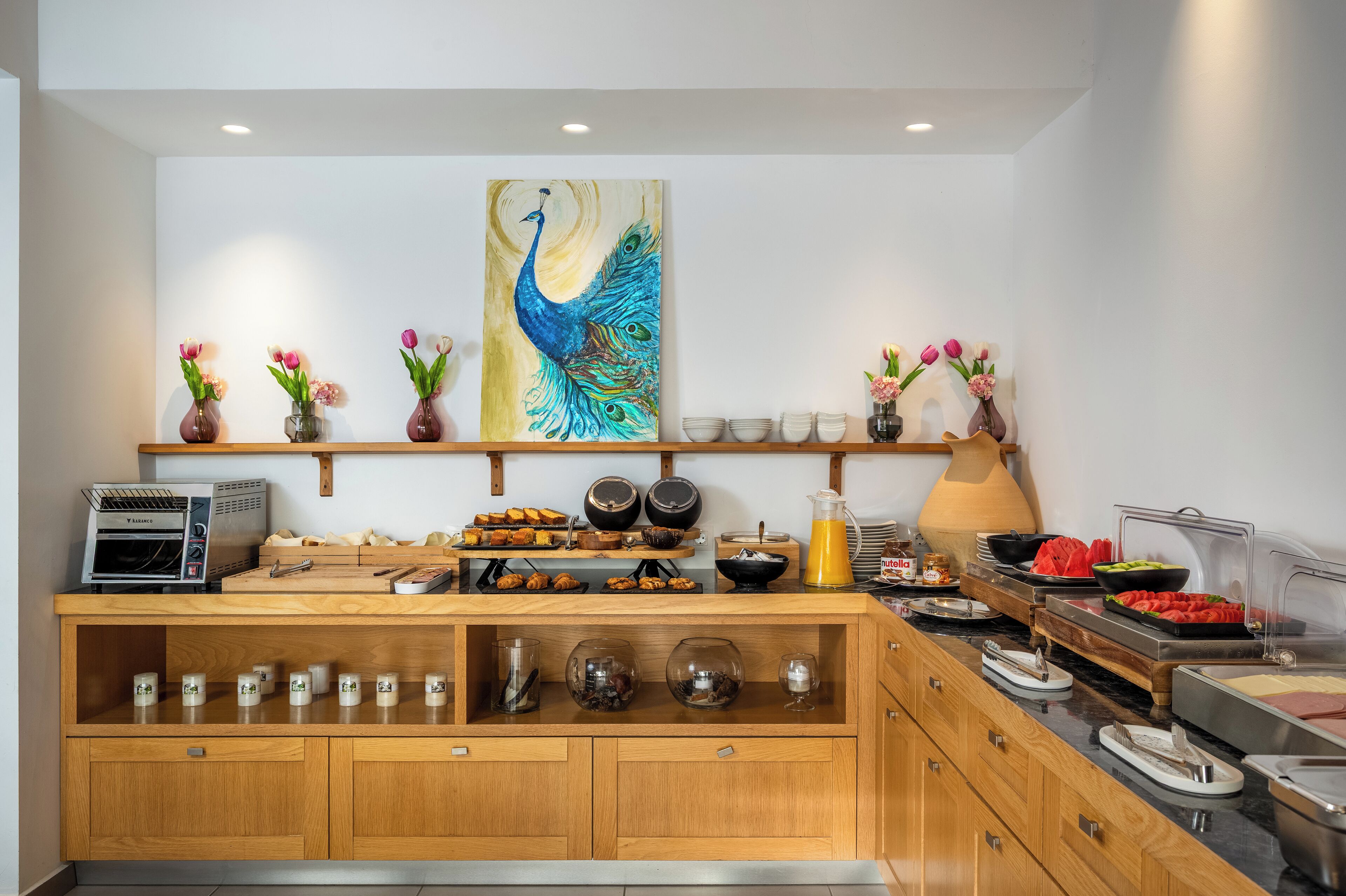 Een buffetopstelling met diverse gerechten, een schilderij van een pauw aan de muur en decoratieve vazen met bloemen.