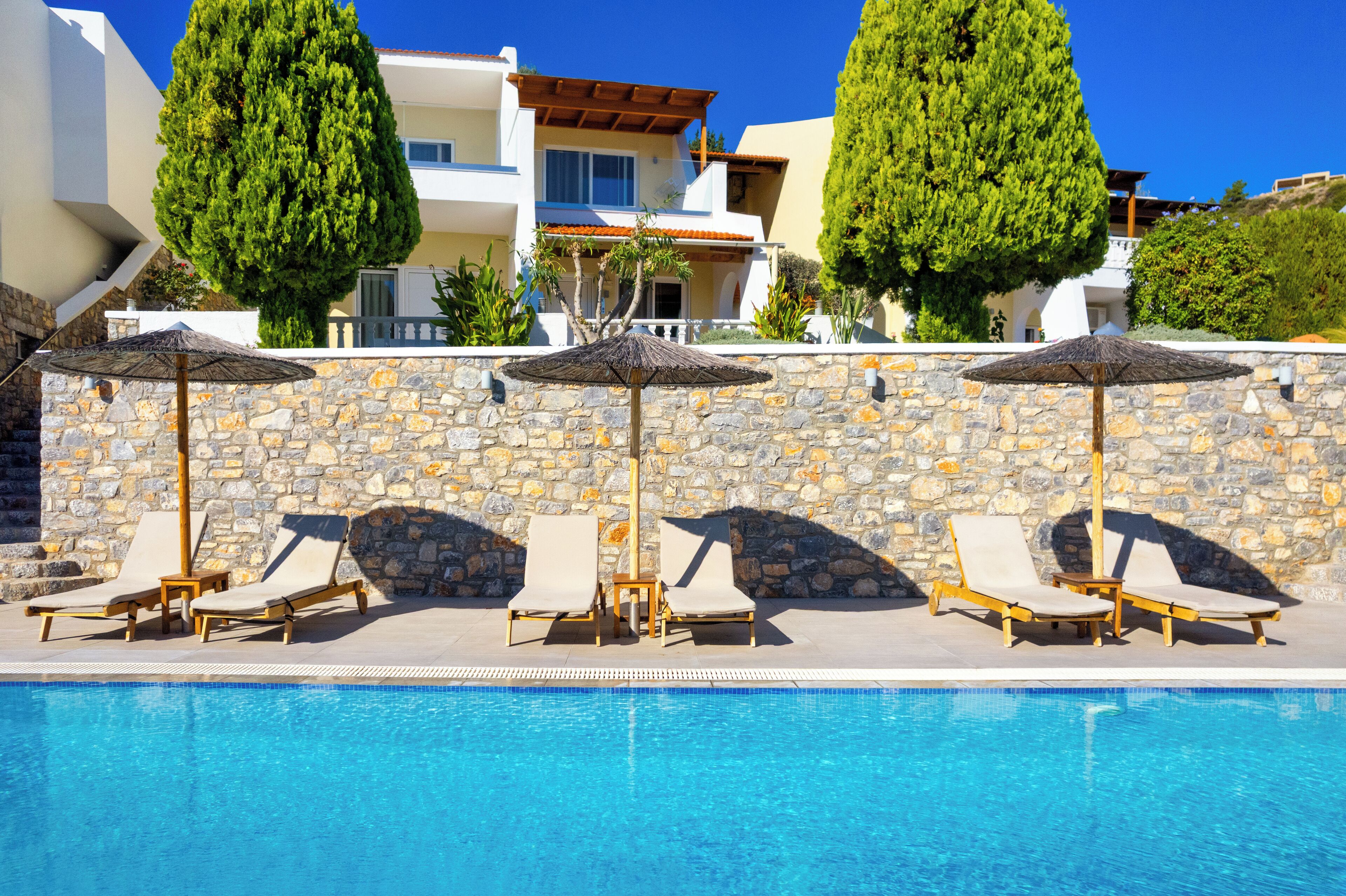 A poolside area with lounge chairs and umbrellas, a stone wall, and a clear blue pool.
