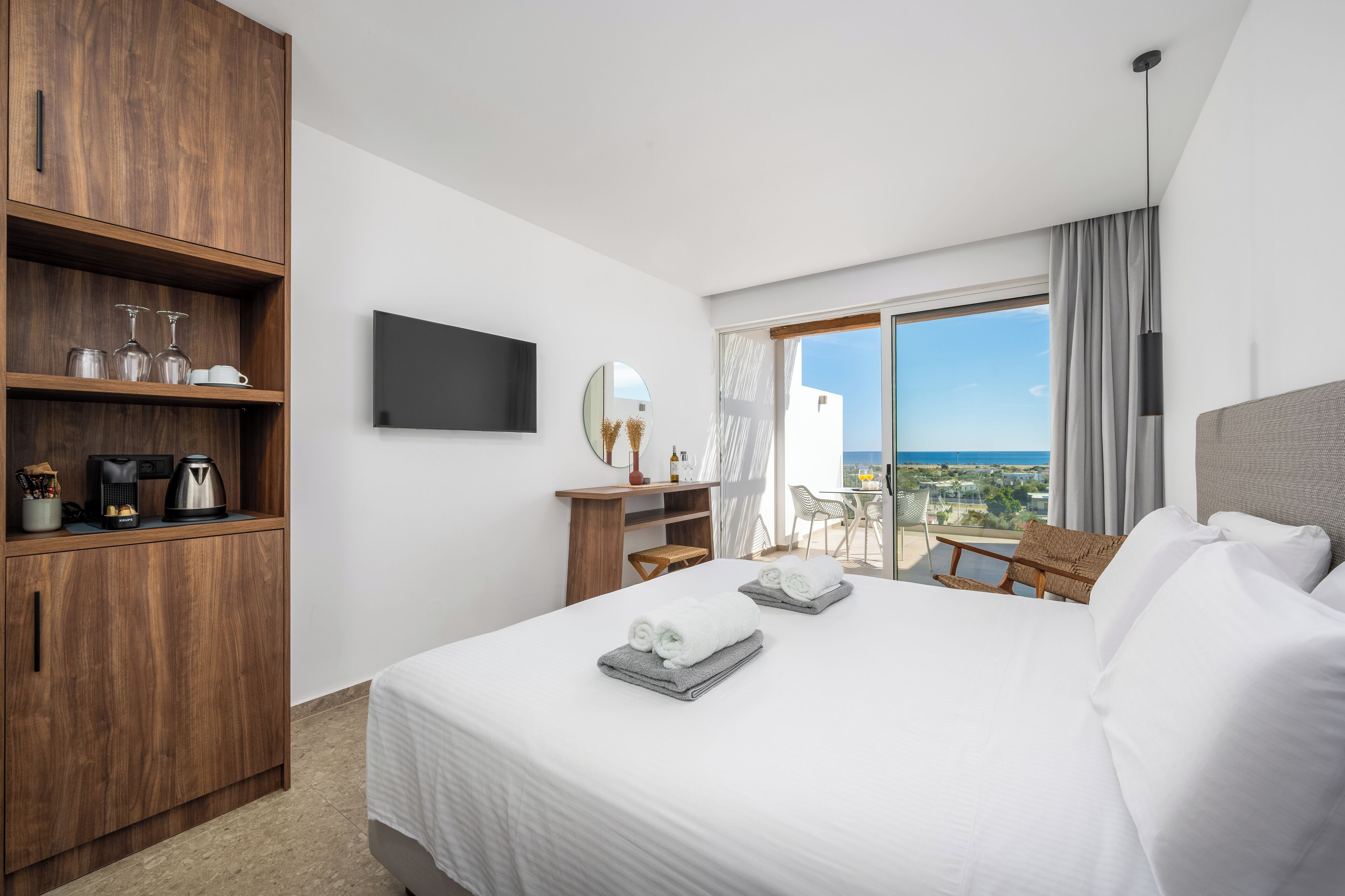 Une chambre d’hôtel comprenant un lit, une armoire en bois, un bureau avec une chaise, un téléviseur à écran plat, un balcon avec vue sur la mer et une fenêtre avec des rideaux.