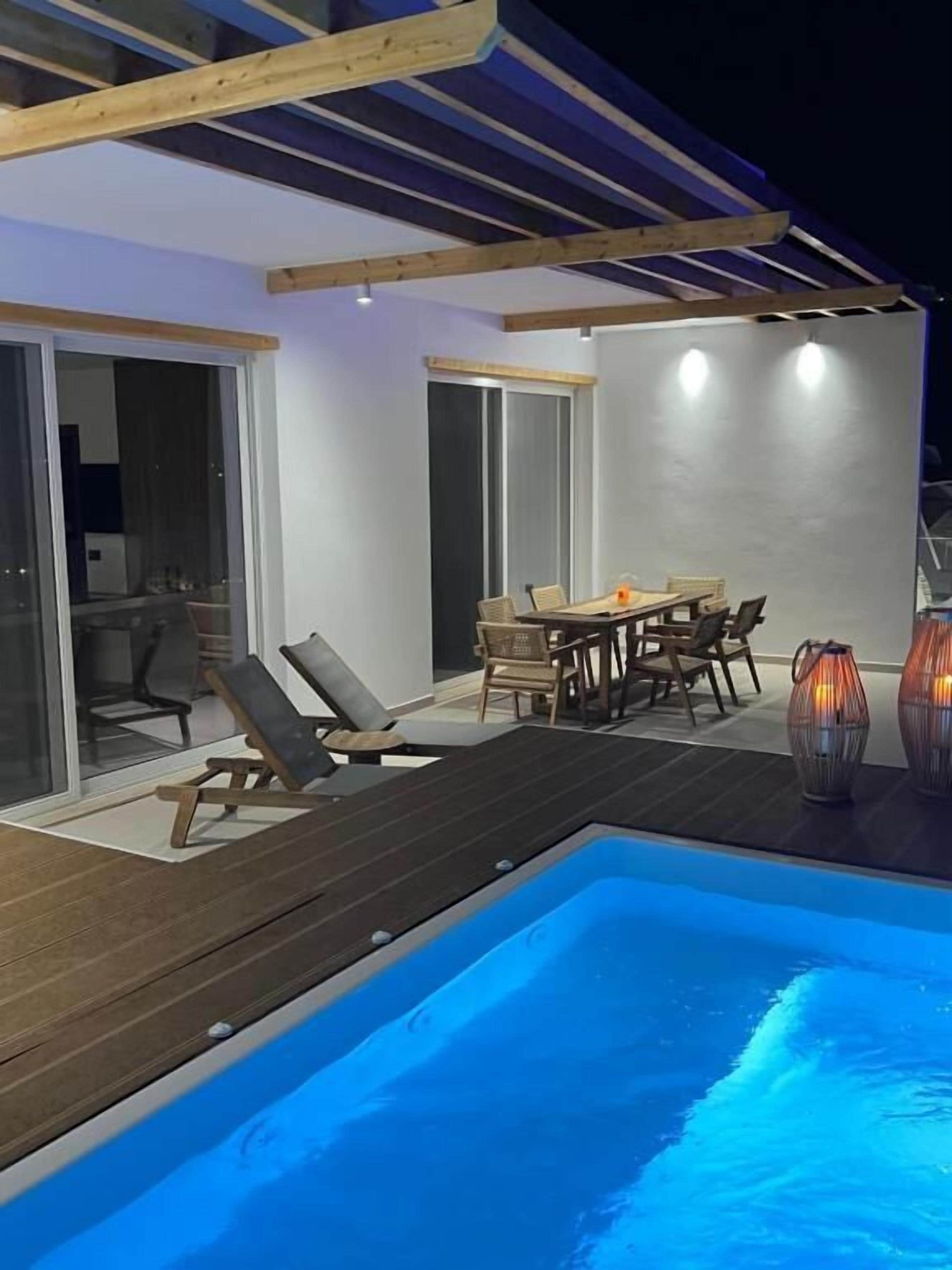 A modern outdoor pool area with a wooden deck, a dining table with chairs, and a lounge chair.