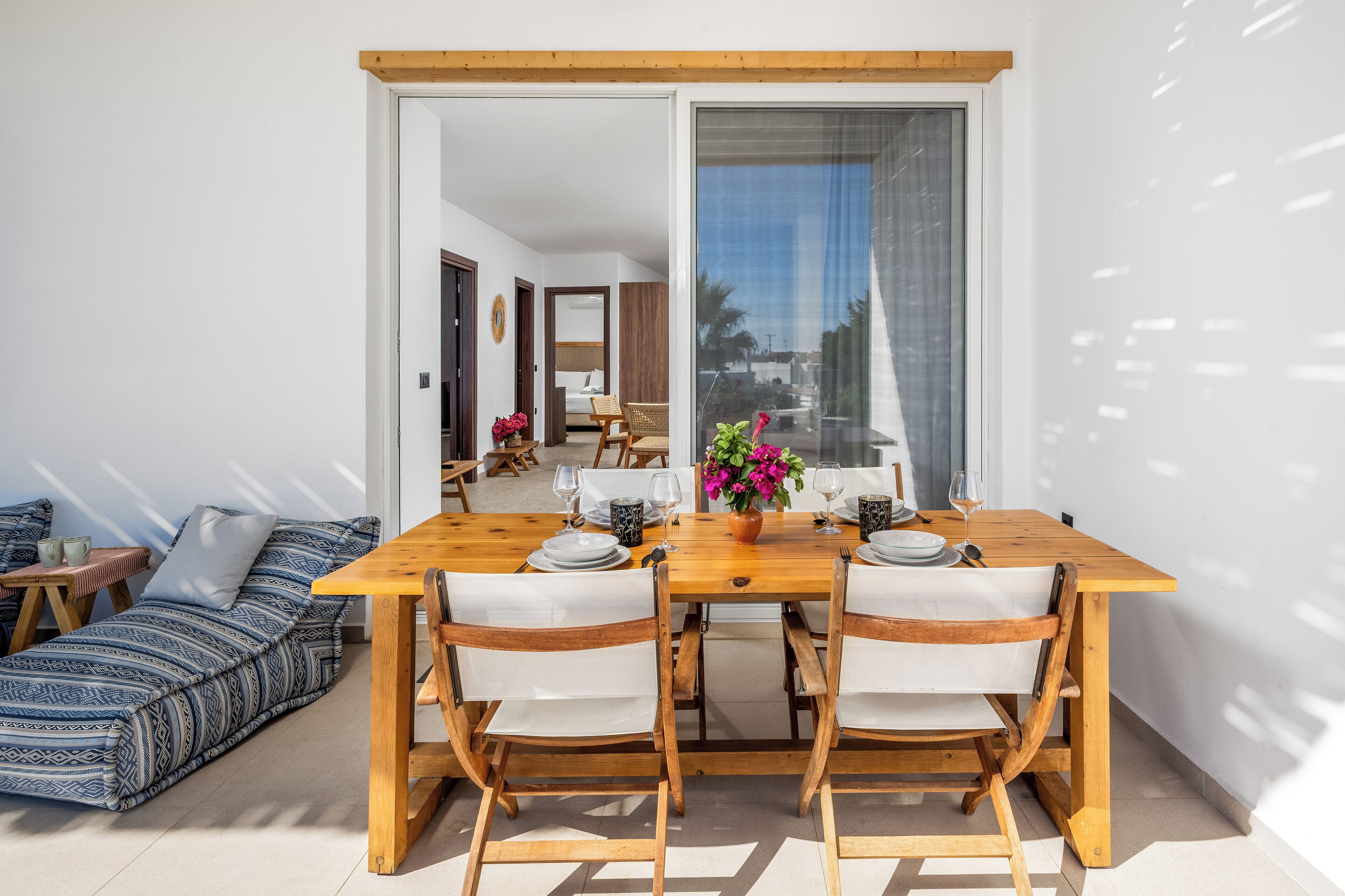 Een eetgedeelte met een houten tafel gedekt voor twee personen, een bank met gestreepte kussens, en een uitzicht door een glazen deur op een woonkamer.