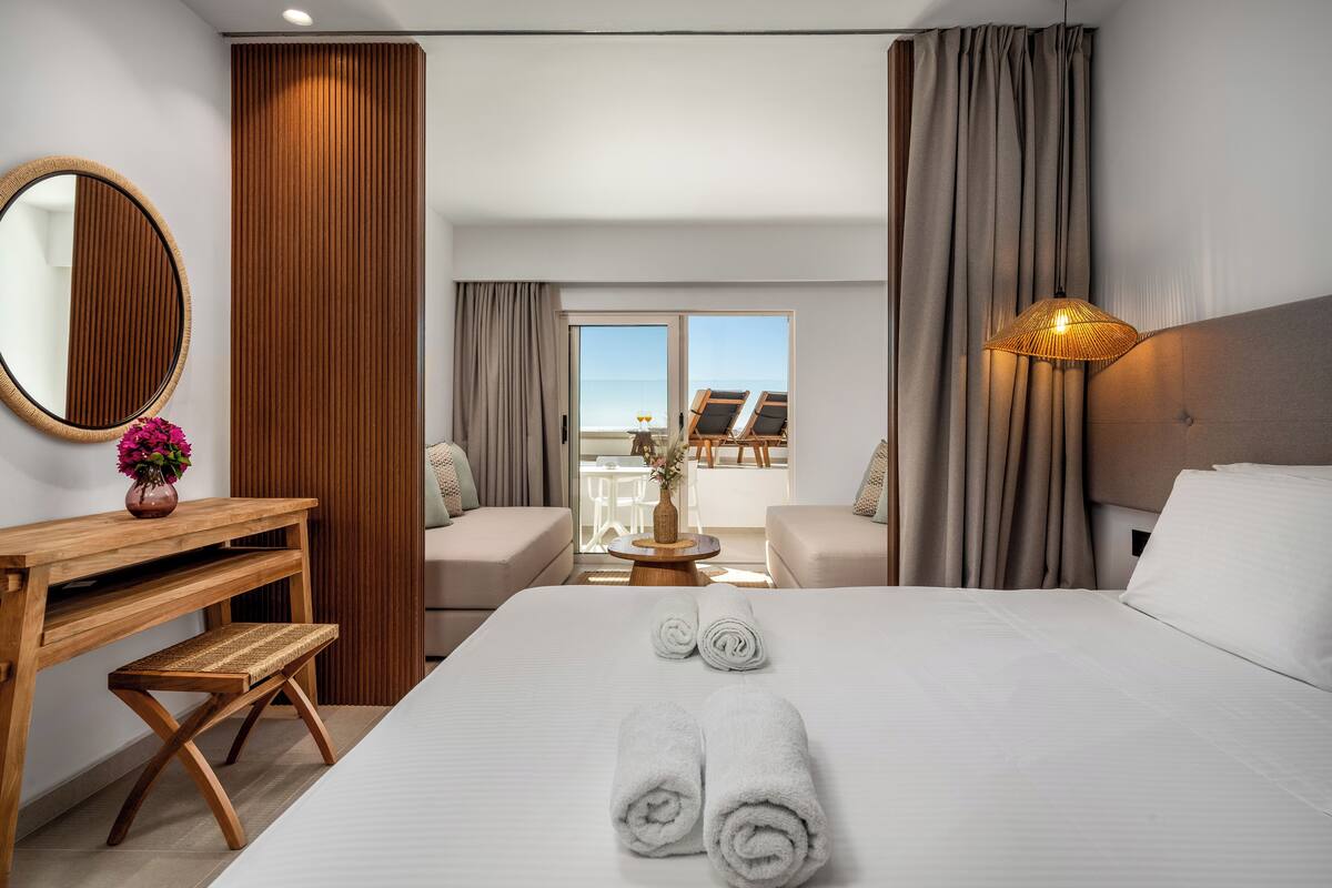 Une chambre d’hôtel avec un grand lit, un bureau en bois, un miroir rond et une vue sur la mer par la fenêtre.