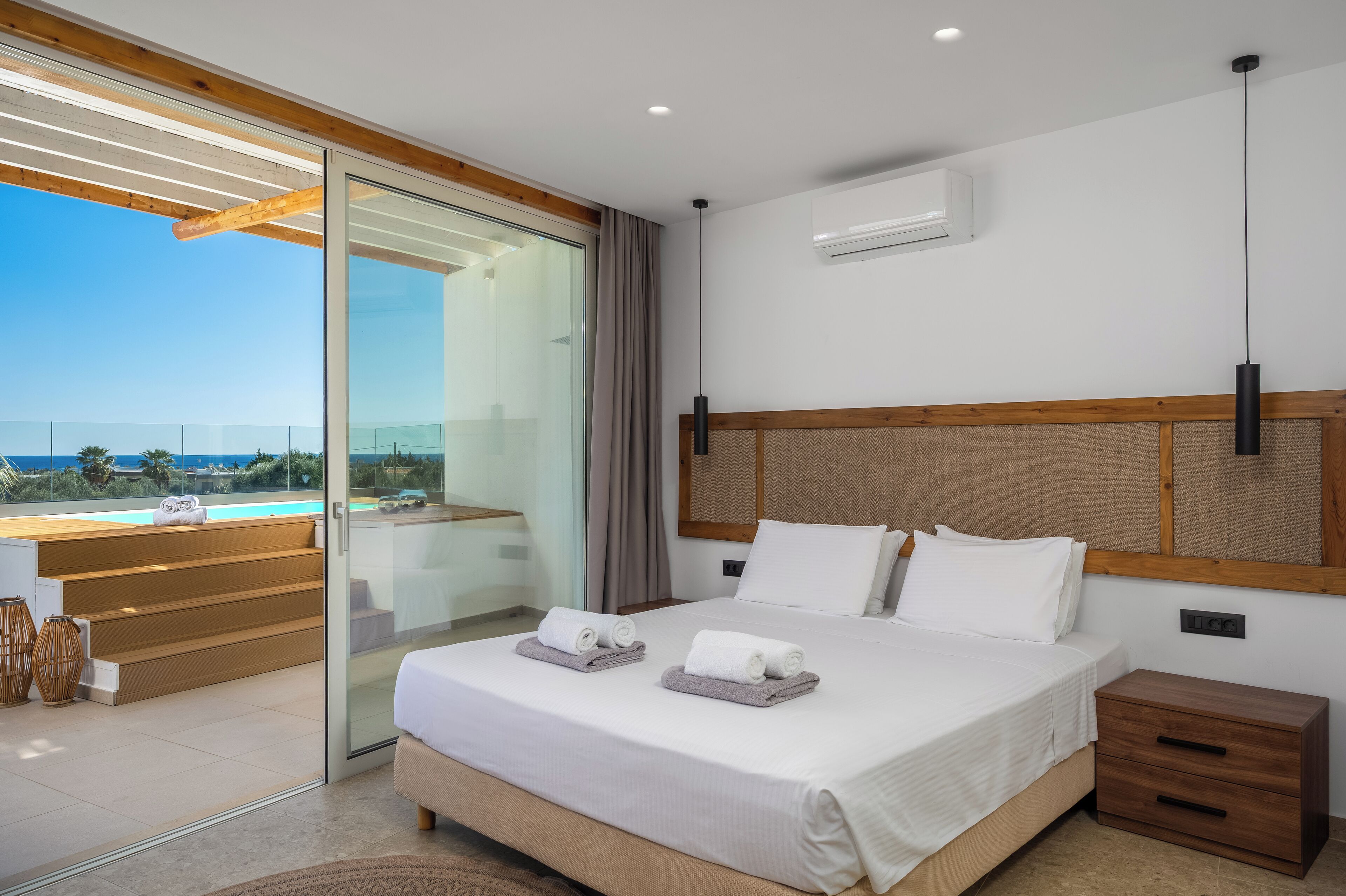 A modern hotel room with a large bed, a wooden headboard, a glass door leading to a balcony with a view of the ocean, and a small wooden nightstand.