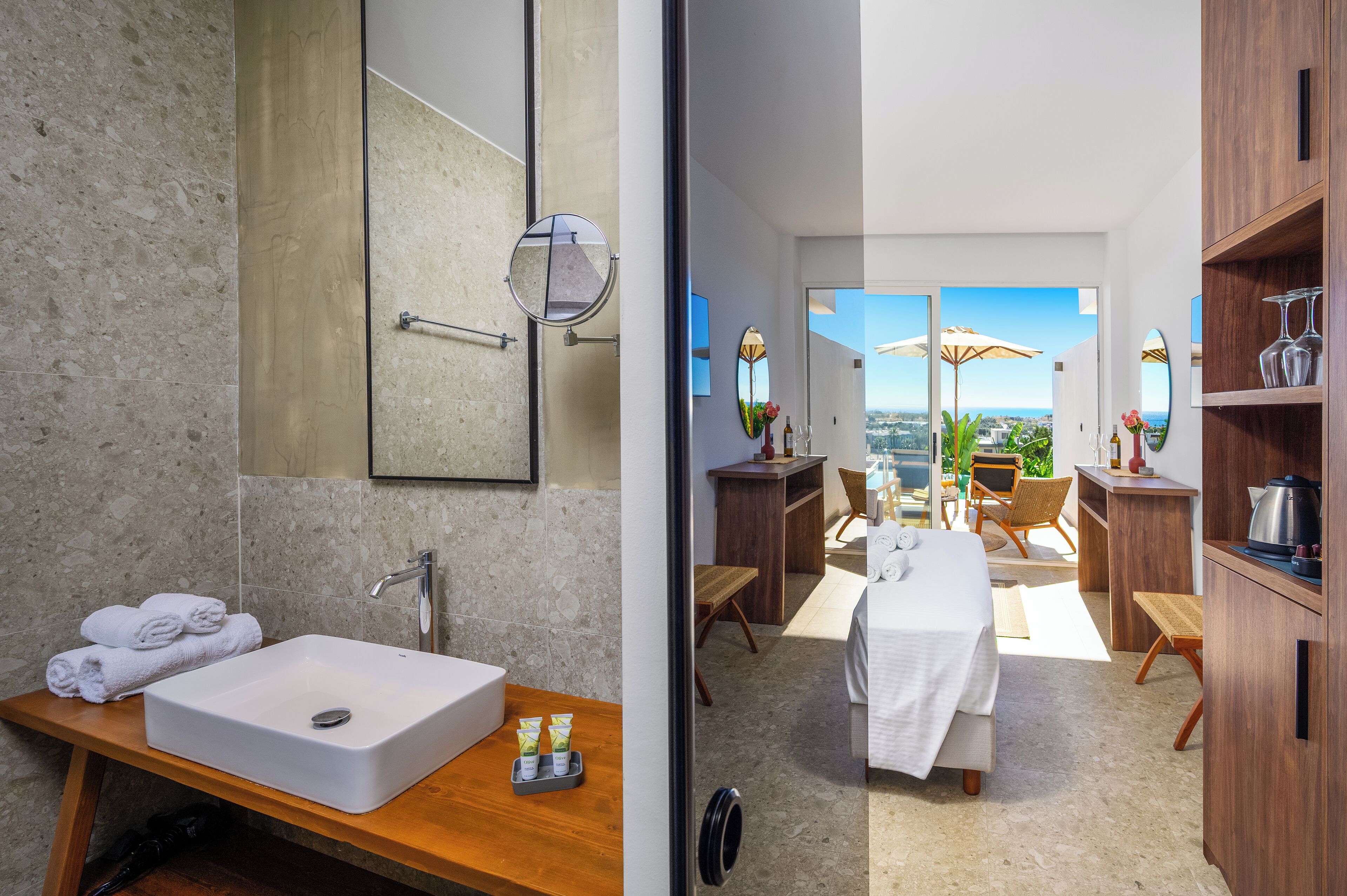 A modern bathroom with a large sink, mirror, and wooden vanity. It leads to a balcony with outdoor seating and a view of the city.