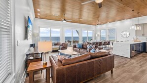 A spacious living area with a large brown leather sofa, wooden flooring, ceiling fans, and a view of the ocean through large windows. - Oytstercatcher: Beachfront, Private Pool, Hot Tub, Sleeps 22! (St. George Island)