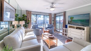 A spacious living room with a large window, a ceiling fan, and a television displaying 'Emerald Beach Properties'.