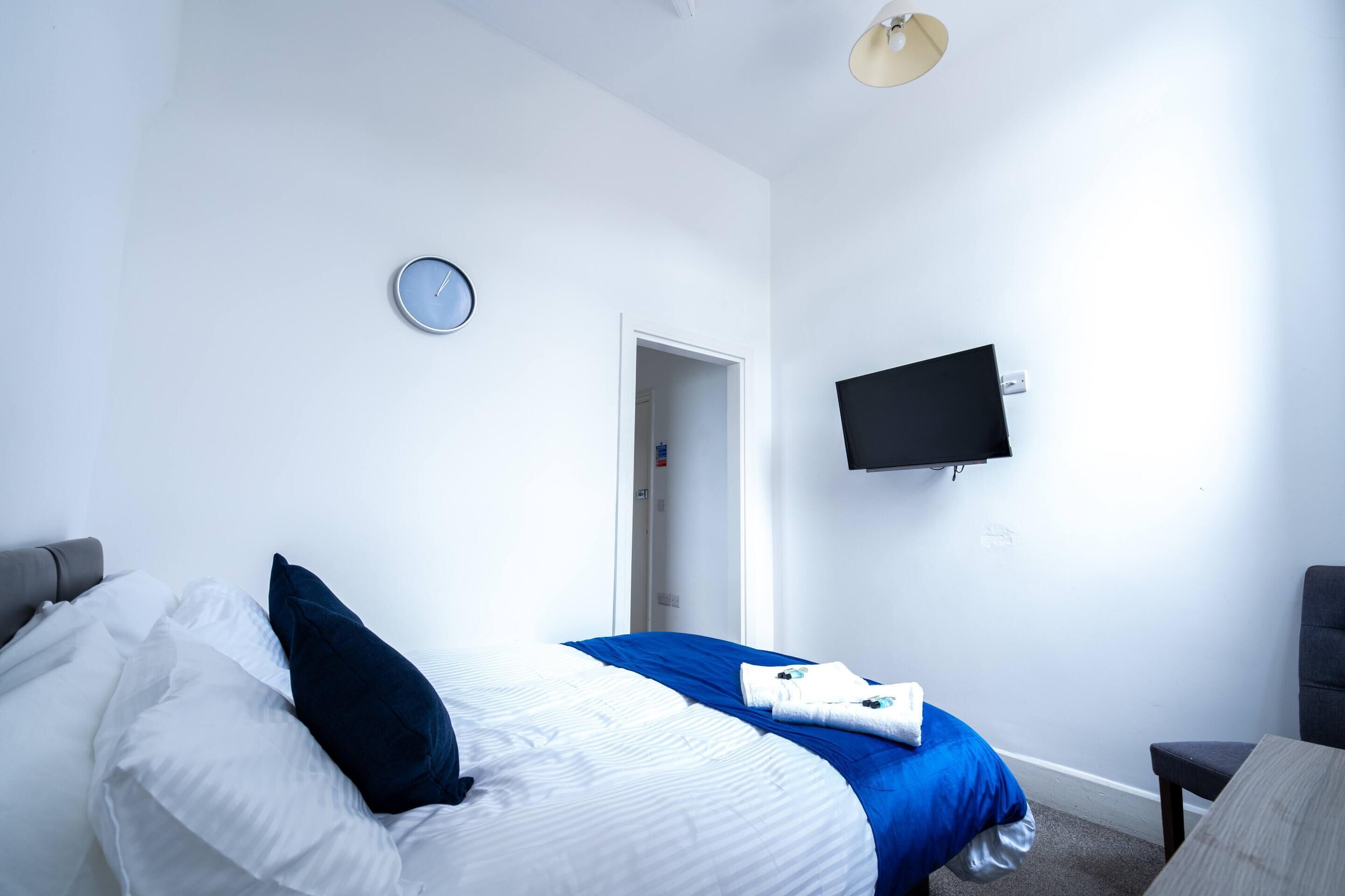 A hotel room with a bed, a TV mounted on the wall, a clock, and a door leading to another room.