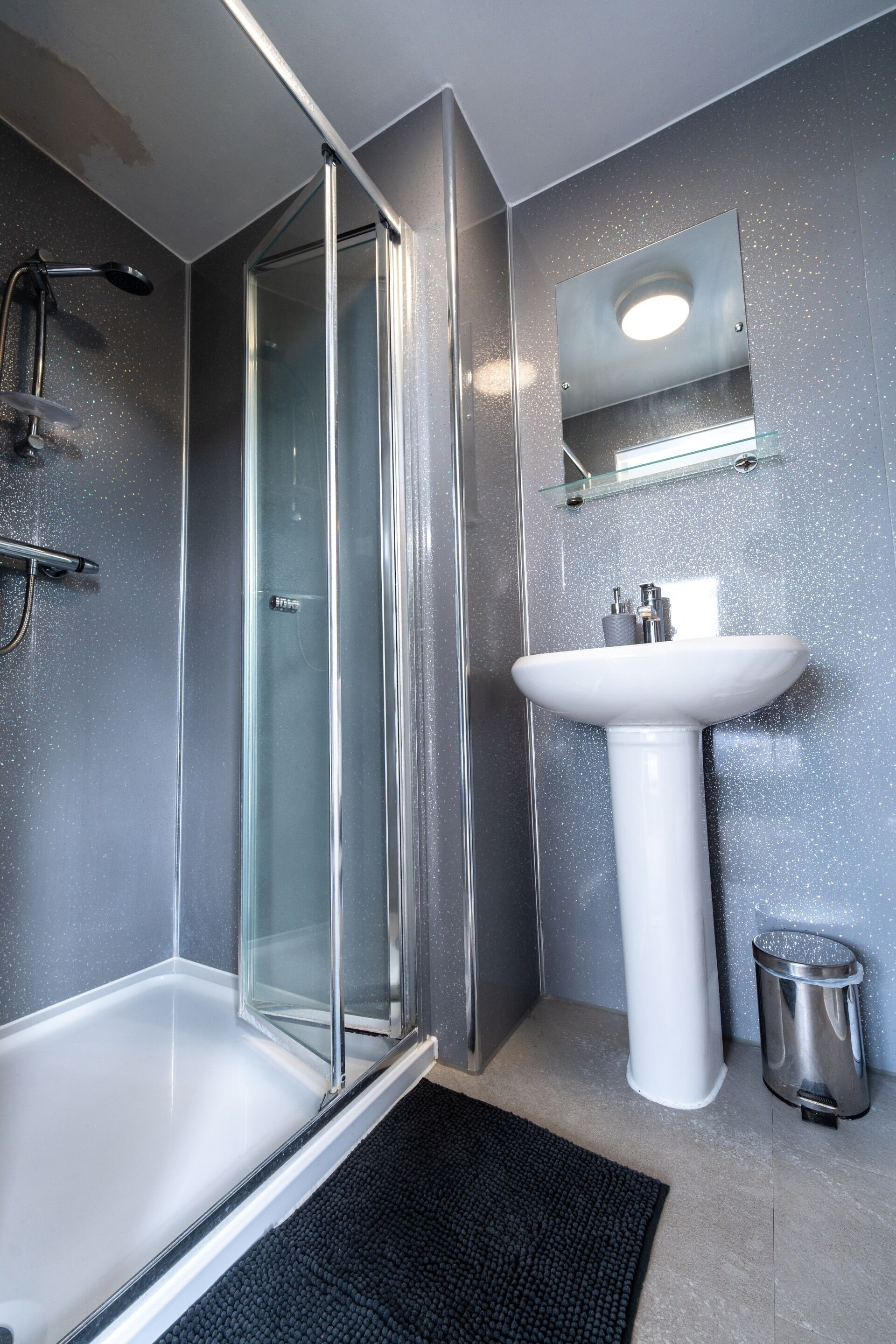 Une salle de bain moderne avec une douche à parois de verre, un lavabo sur pied blanc et une petite poubelle.