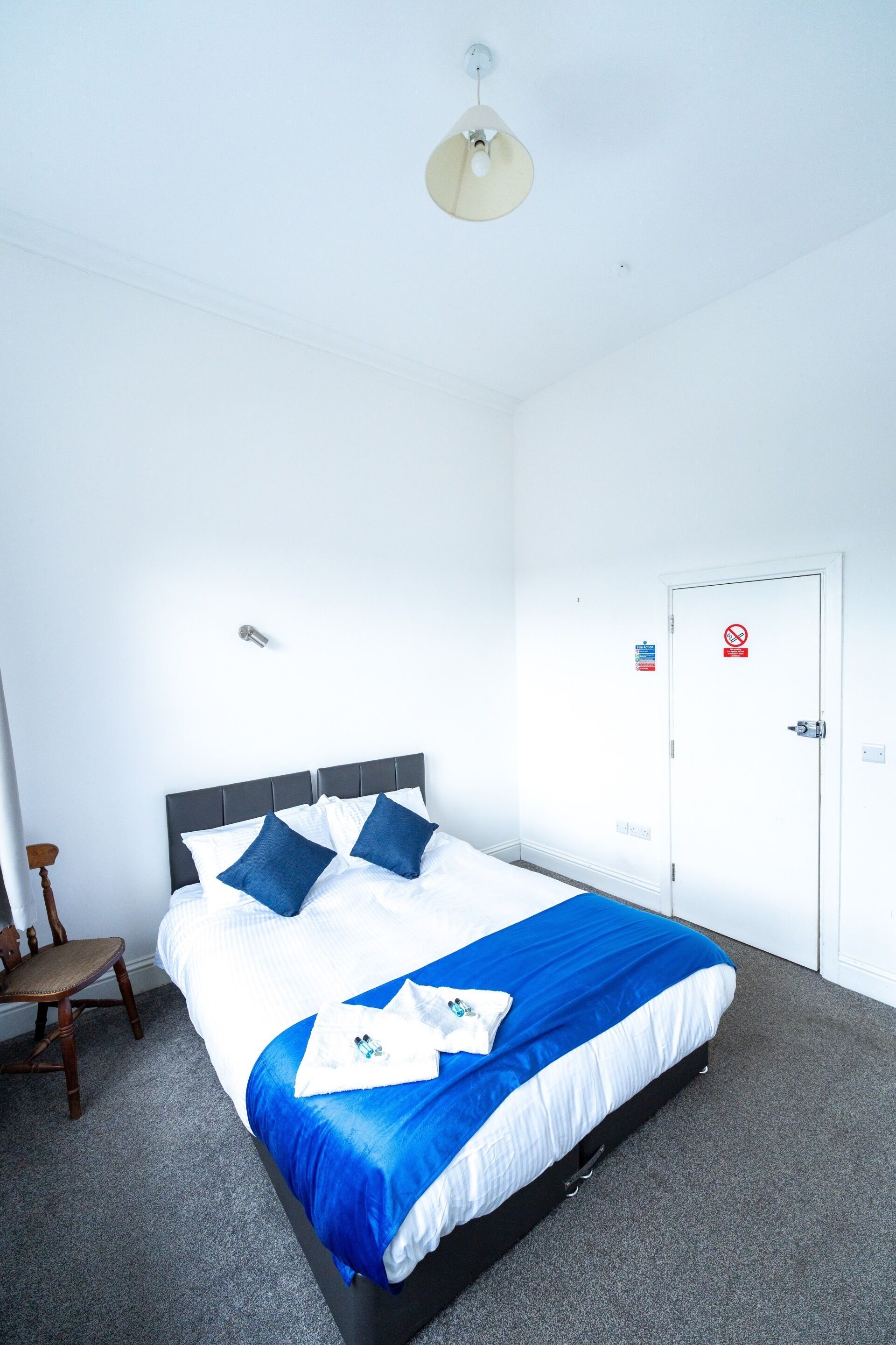 A neatly made bed with blue and white bedding, a wooden chair, a white door with a no smoking sign, and a ceiling light fixture.
