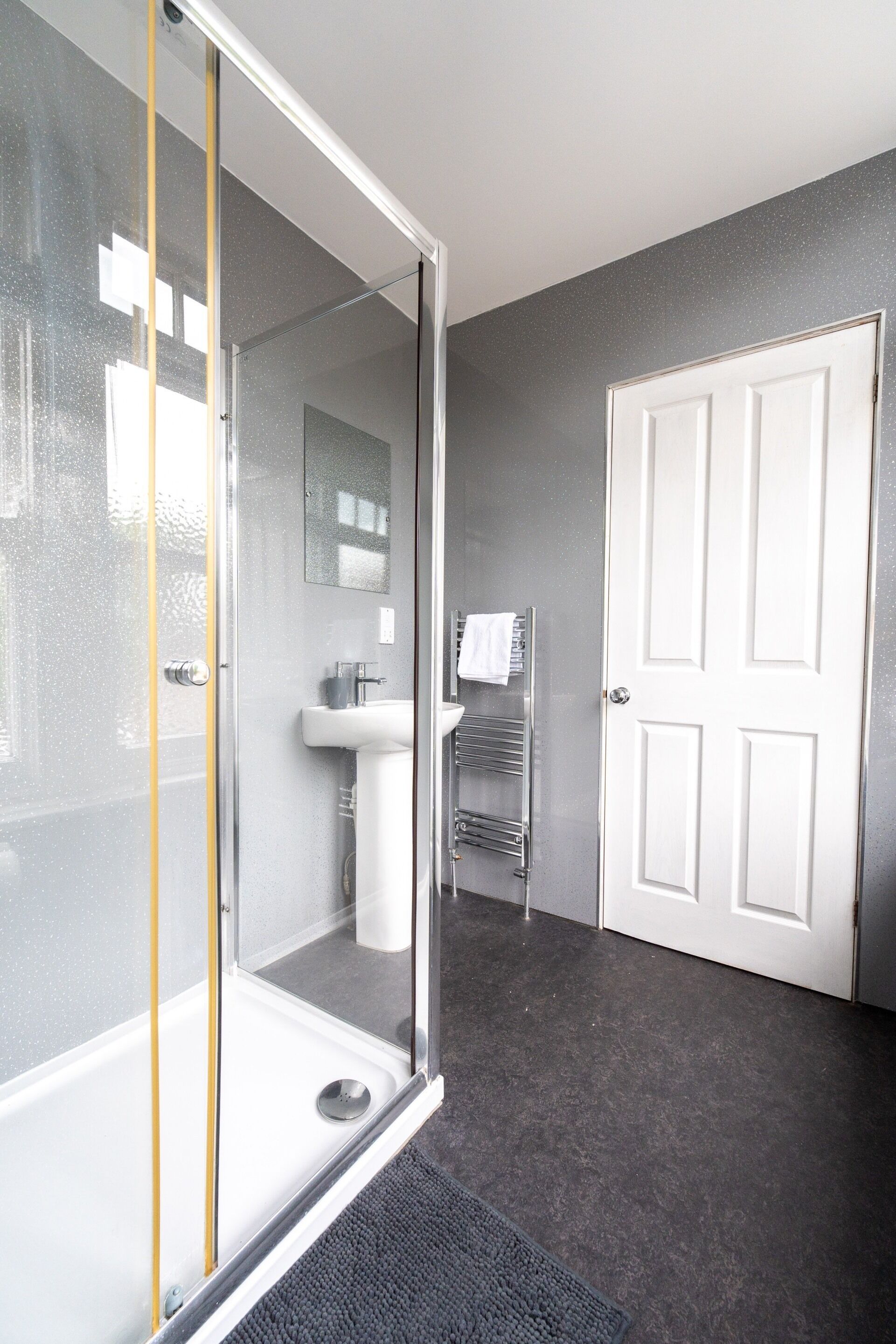 A bathroom with a glass shower enclosure, a white sink, a towel rack, and a white door.