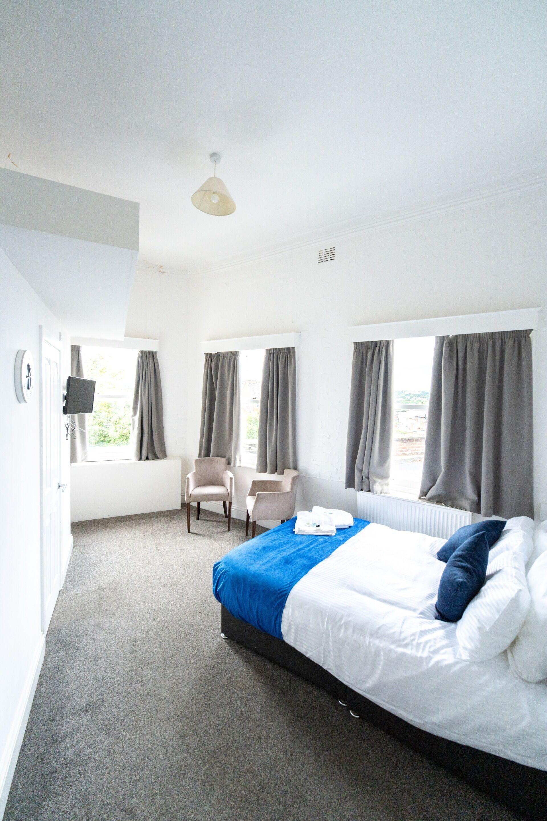 A hotel room with a bed, two chairs, a desk, a TV, and large windows with curtains.