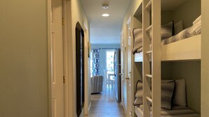 A narrow hallway with wooden flooring leading to a room with bunk beds and a television mounted on the wall. - Ocean View - Ocean Villas B215-Professionally Managed by HHIB&T Vacation Rentals (Hilton Head Island)