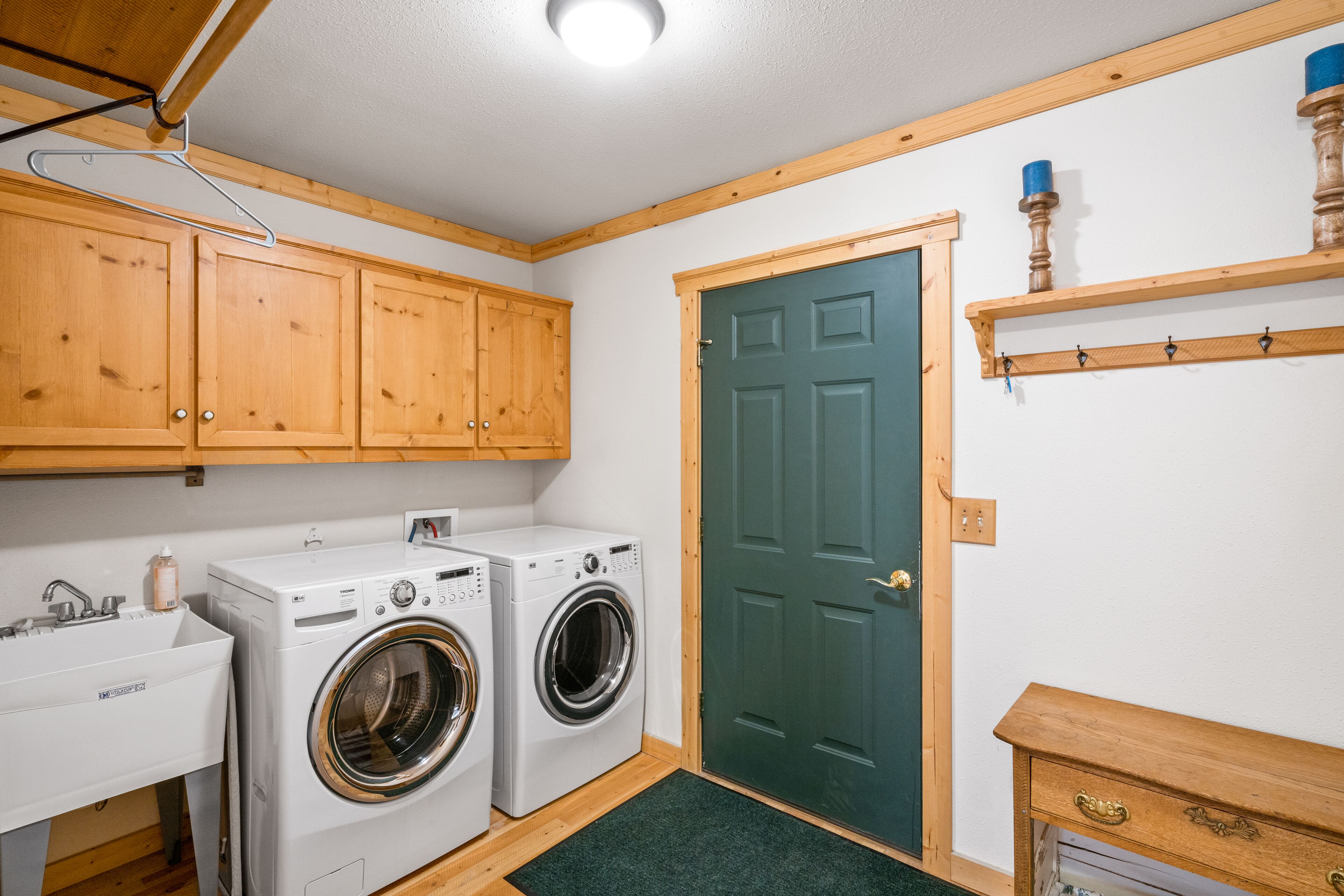 A compact laundry room with two washing machines, a sink, wooden cabinets, a green door, and a shelf with hooks.