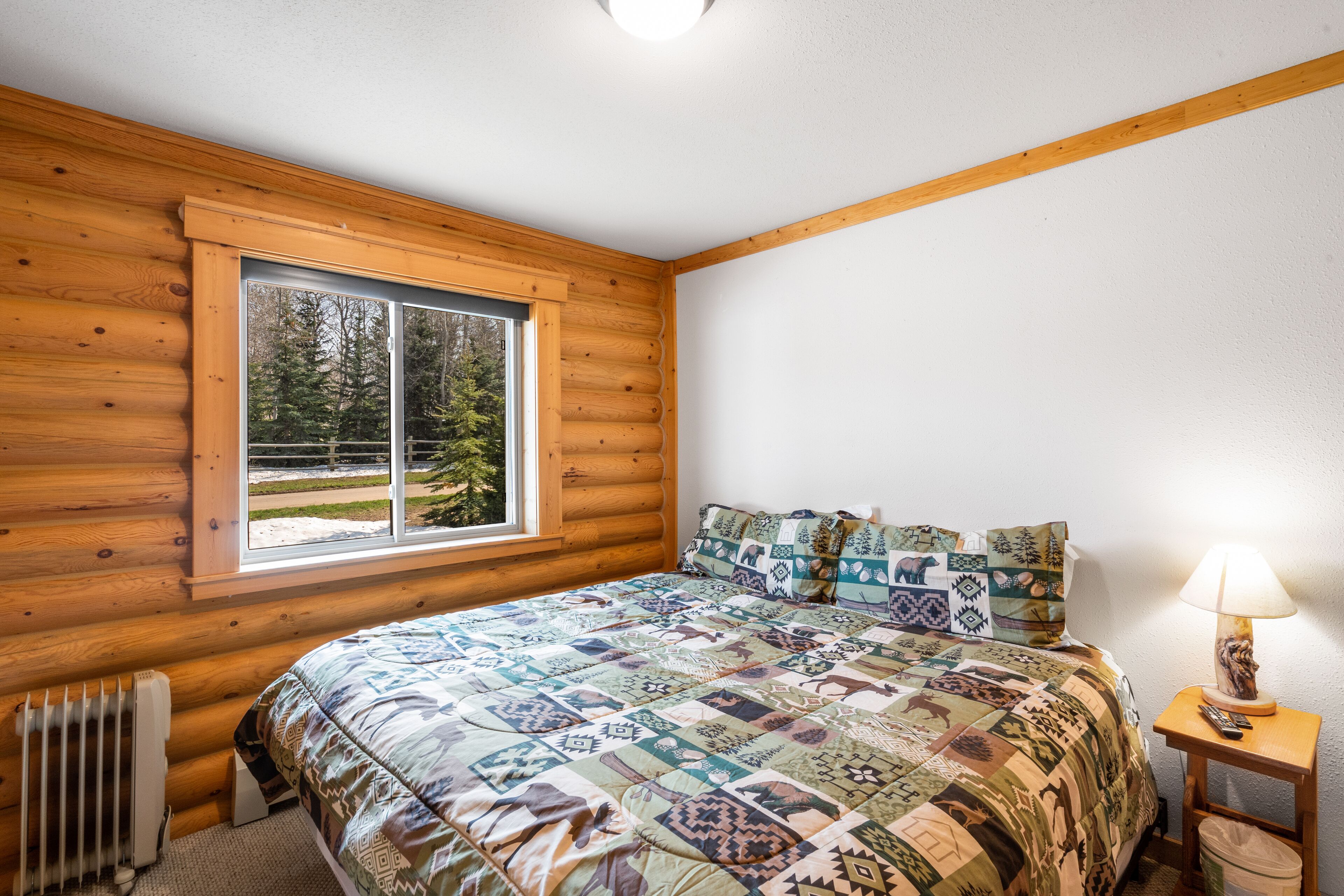 Un dormitorio tipo cabaña de madera con una cama grande, una ventana con vistas a árboles, un radiador y una mesita de noche con una lámpara.
