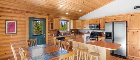A wooden cabin interior with a kitchen, dining area, and a view of the forest.