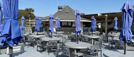 An outdoor seating area with round tables and chairs, covered by blue umbrellas, and a building in the background.