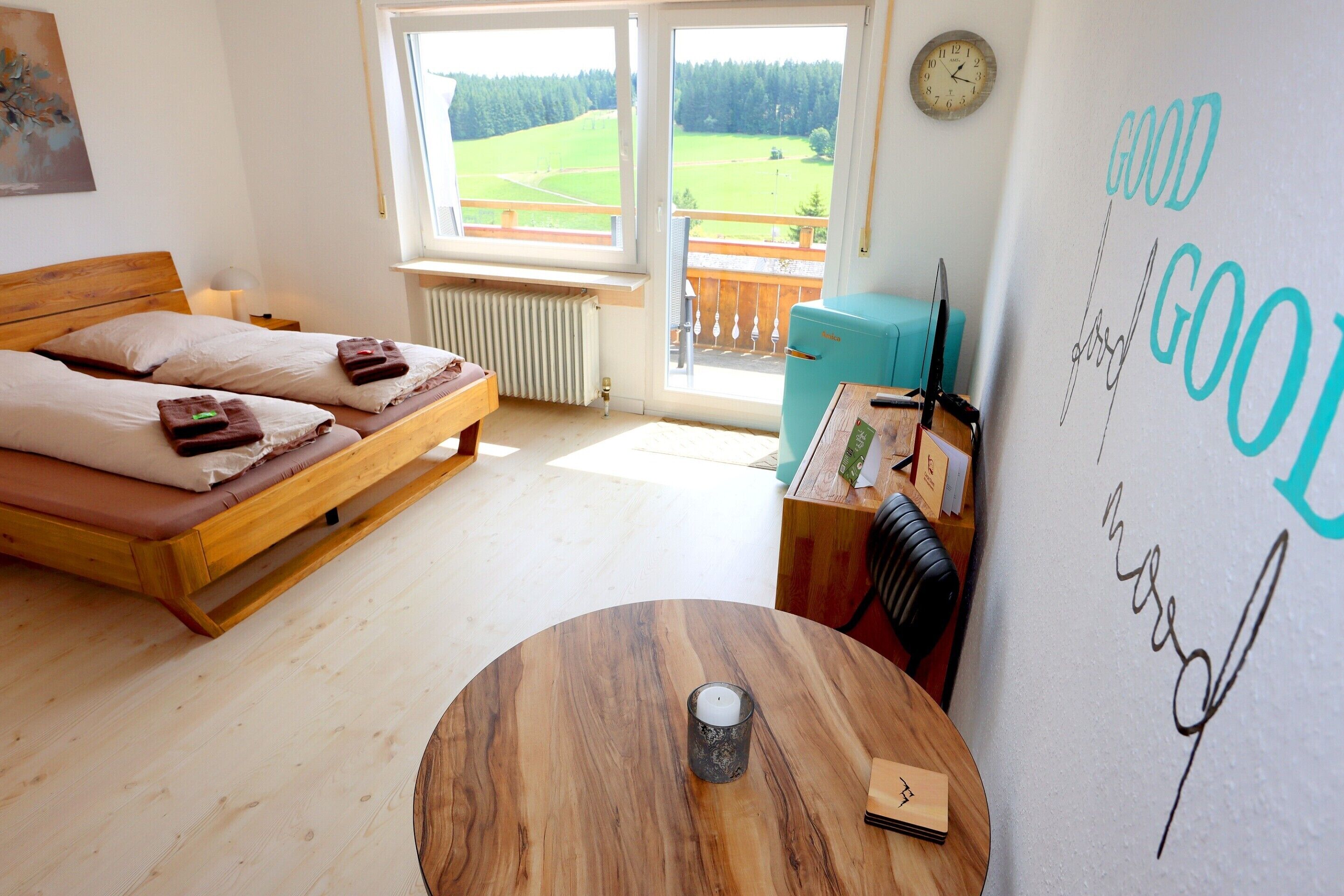 Een slaapkamer met een bed, een ronde houten tafel, een wandklok en uitzicht op een groen veld door een raam.