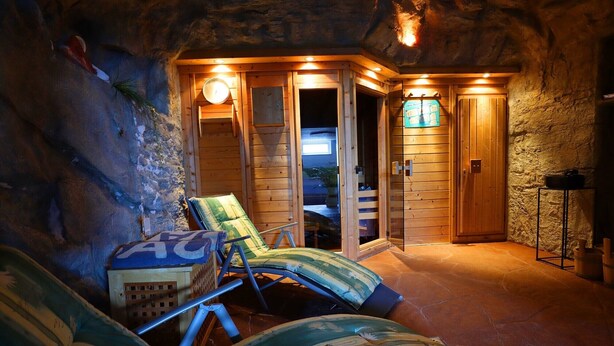 Une salle de spa chaleureuse et rustique, avec des murs en bois et un sol en pierre. On y trouve deux fauteuils inclinables dotés de coussins à rayures. Une porte en bois donne accès à une zone douche, et une petite table avec une lampe est visible.