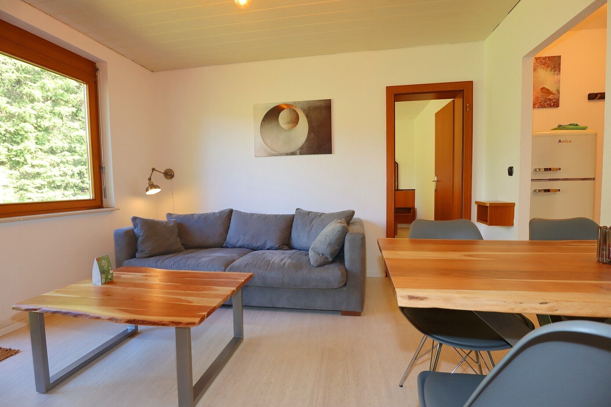 A modern living room with a grey sofa, a wooden coffee table, and a dining table with chairs. There is a painting on the wall and a window with a view of trees.