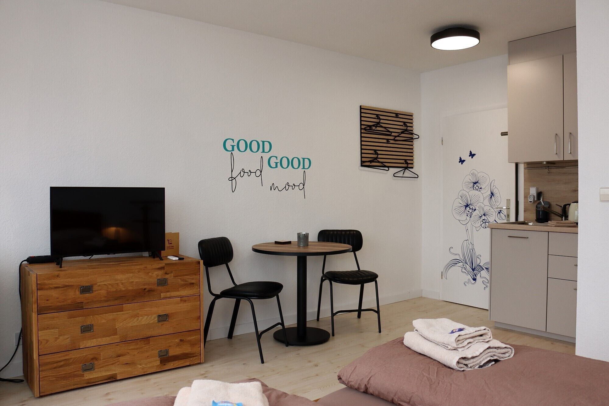 Una habitación compacta con un aparador de madera, un televisor, una pequeña mesa de comedor con sillas, una kitchenette y una pared con un adhesivo que dice 'GOOD food GOOD mood'.
