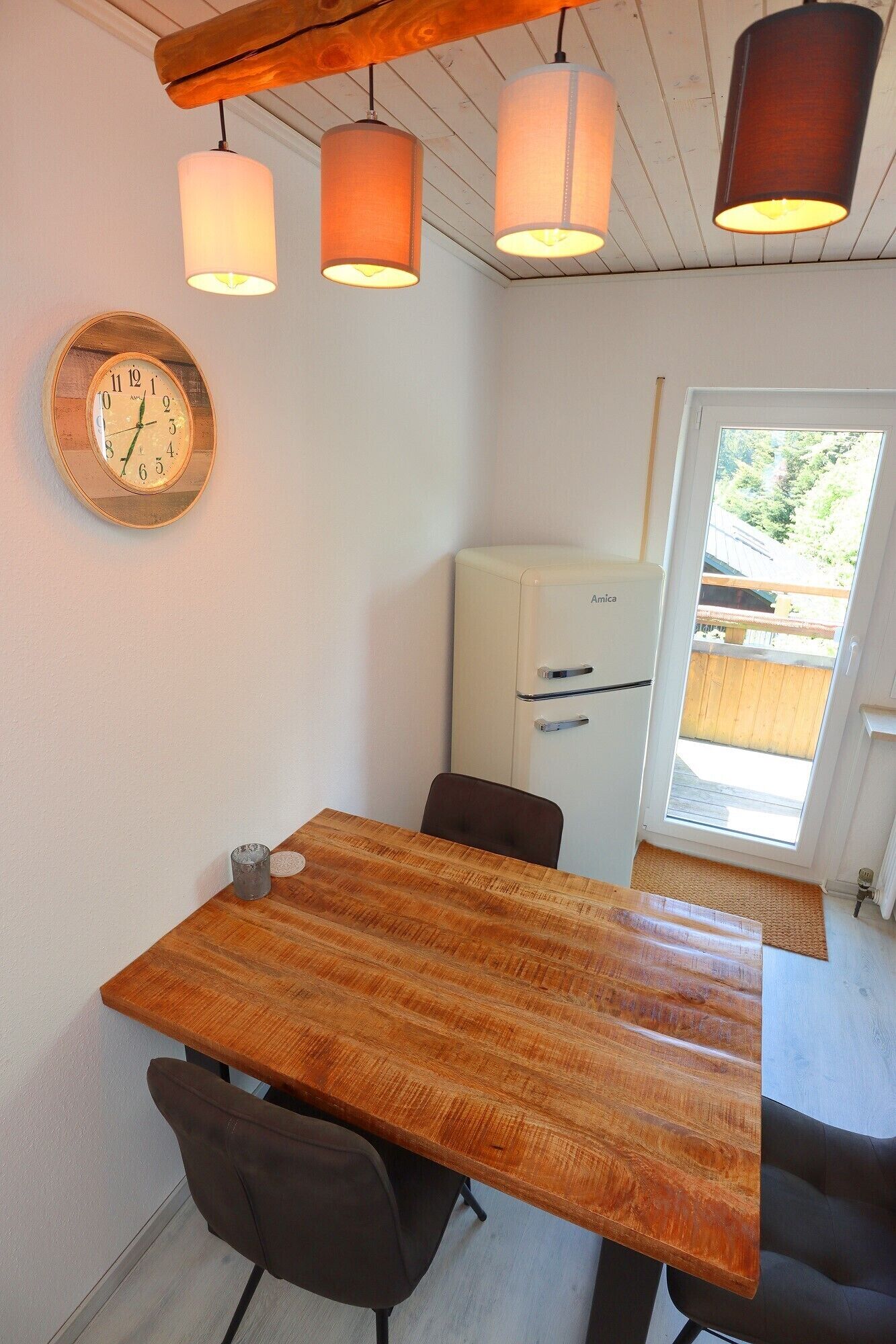 A dining area with a wooden table, chairs, pendant lights, a clock, and a refrigerator.