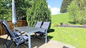 En udendørs terrasse med et metalbord og to stole, omgivet af et trædæk og en græsplæne med træer.