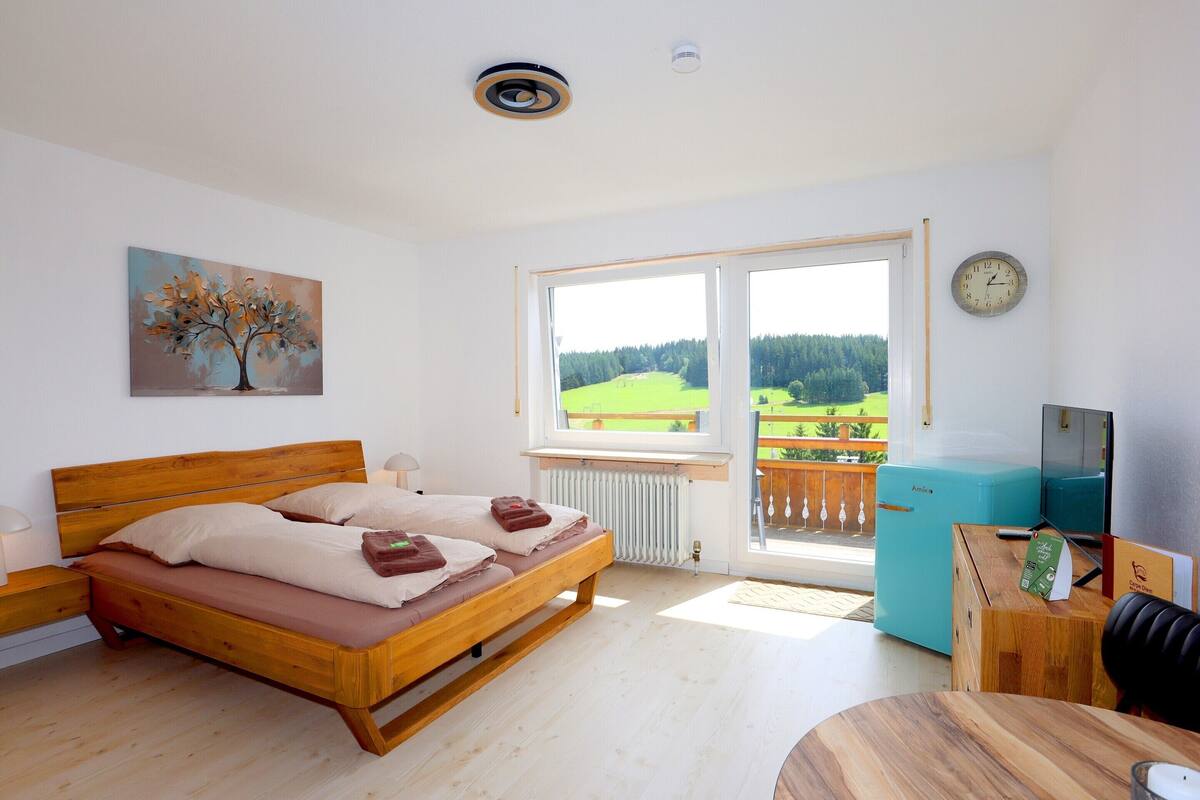 A bedroom with a wooden bed, a painting of a tree, a view of a green landscape through the window, and a clock on the wall.