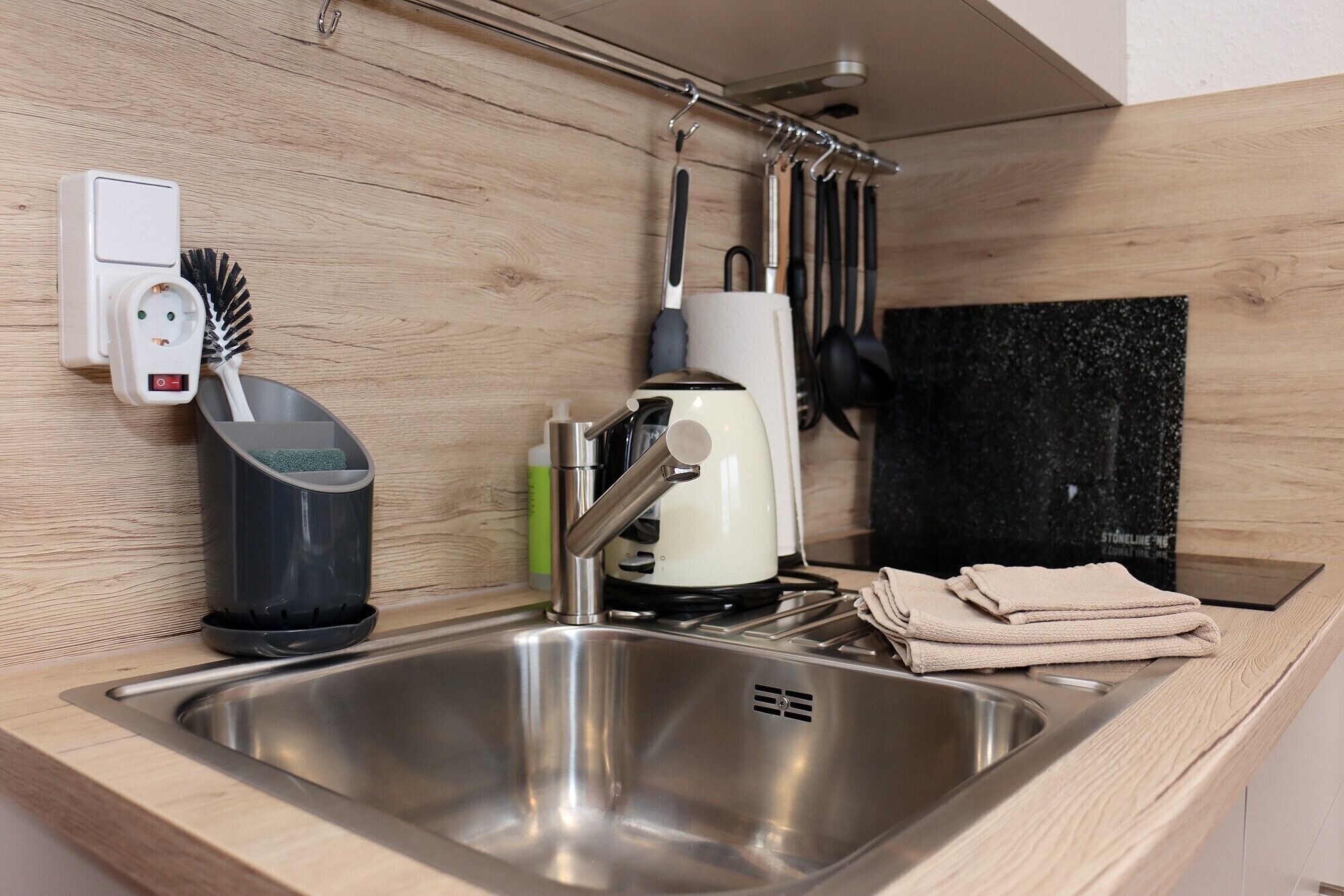 A modern kitchen with a stainless steel sink, a kettle, and a coffee maker. There is a power outlet and a set of utensils hanging on the wall.